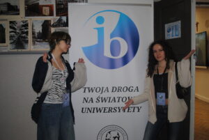 Dwie uczennice stoją przy banerze IB z napisem „Twoja droga na światowe uniwersytety”, prezentując go gestem rąk.