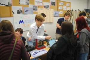 Stoisko szkolne z plakatami „Maths SL & HL” i „Taniec funkcji”. Uczniowie rozmawiają i prezentują materiały edukacyjne.