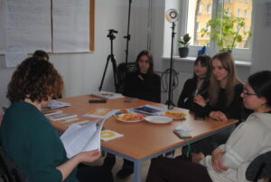 Dyskusja przy stole – nauczycielka czyta dokument, a kilka uczennic słucha. Na stole leżą wydruki, telefony i cukierki.