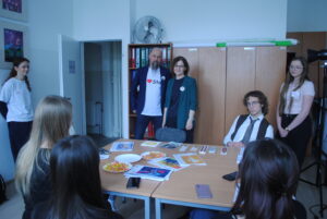 Spotkanie przy stole – uczniowie siedzą wokół, a nauczyciel i nauczycielka stoją na końcu sali. Na stole materiały, kartki i słodycze.