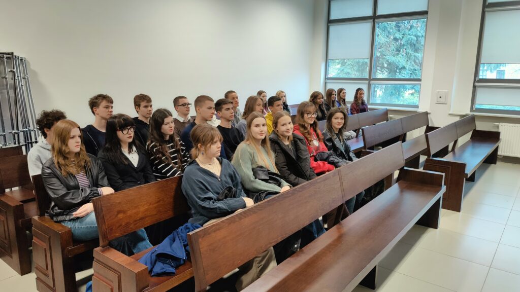 Sala przypominająca szkolną lub sądową, z drewnianymi ławami ustawionymi w rzędach. Na ławach siedzi grupa kilkunastu młodych osób (uczniów), ubranych w codzienne stroje – swetry, bluzy, koszule. Większość patrzy w jednym kierunku, z neutralnymi lub lekko skupionymi wyrazami twarzy. Po prawej stronie widoczne są duże okna wpuszczające światło dzienne, za którymi widać drzewa. Atmosfera jest spokojna i uporządkowana.