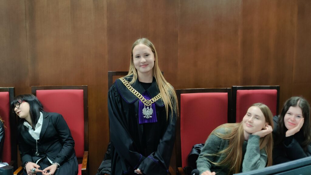 Wnętrze sali przypominającej sąd, z drewnianą ścianą w tle i czerwonymi, wysokimi krzesłami. Na środku stoi młoda kobieta w todze sędziowskiej z ozdobnym łańcuchem z orłem na piersi. Ma długie, jasne włosy i delikatnie się uśmiecha, patrząc w stronę aparatu. Po jej lewej stronie siedzi kobieta w czarnym stroju, z głową przechyloną na bok i zamkniętymi oczami, jakby była zmęczona lub przysypiała. Po prawej stronie siedzą dwie młode kobiety, oparte o ręce, patrzące z lekkim uśmiechem w stronę osoby stojącej.