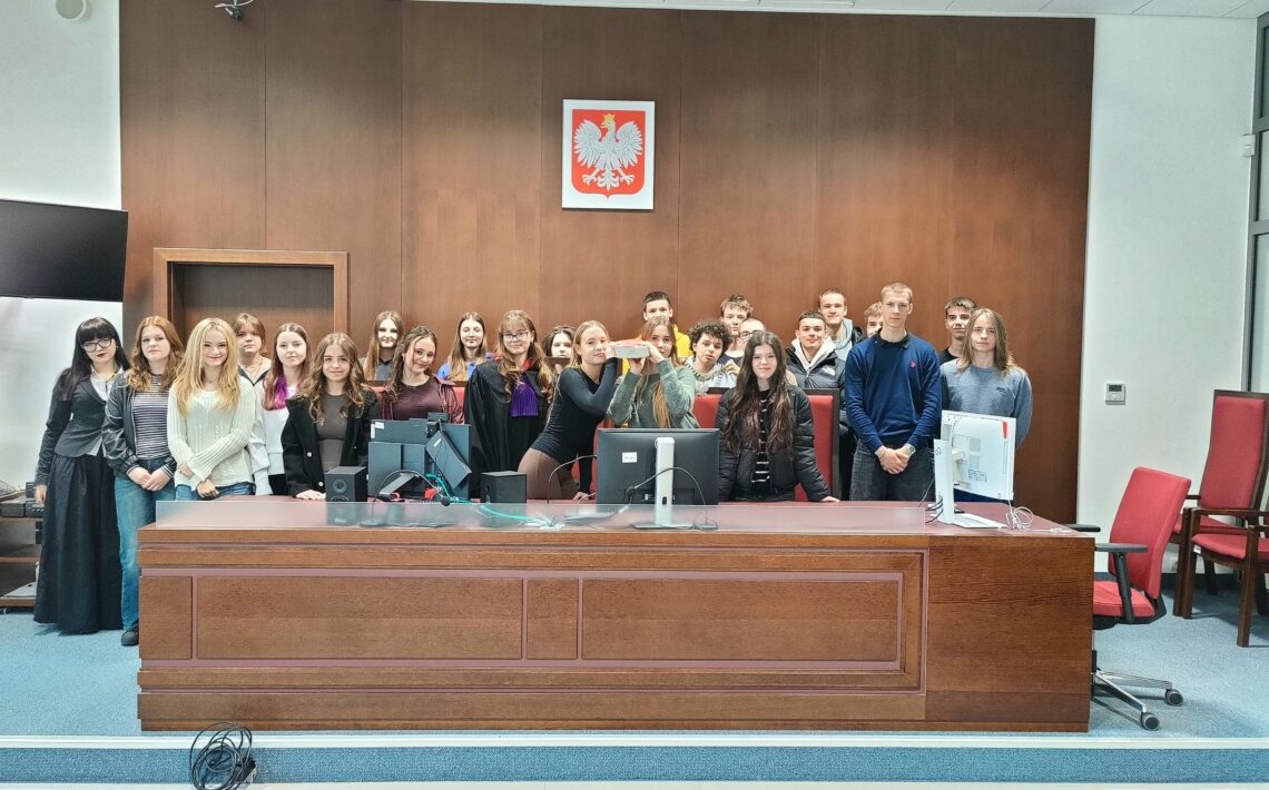 Duża grupa młodych osób stoi za wysokim, drewnianym stołem sędziowskim w sali sądowej. Na ścianie nad nimi widnieje godło Polski – biały orzeł na czerwonym tle. Na stole znajdują się monitory komputerowe, klawiatury i okablowanie. W centrum kilka osób trzyma przedmiot przypominający pudełko lub rekwizyt, prezentując go. Uczestnicy stoją blisko siebie, część uśmiecha się do zdjęcia. Po bokach widać czerwone krzesła oraz elementy wyposażenia sali.