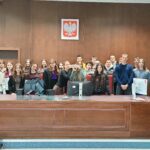 Duża grupa młodych osób stoi za wysokim, drewnianym stołem sędziowskim w sali sądowej. Na ścianie nad nimi widnieje godło Polski – biały orzeł na czerwonym tle. Na stole znajdują się monitory komputerowe, klawiatury i okablowanie. W centrum kilka osób trzyma przedmiot przypominający pudełko lub rekwizyt, prezentując go. Uczestnicy stoją blisko siebie, część uśmiecha się do zdjęcia. Po bokach widać czerwone krzesła oraz elementy wyposażenia sali.