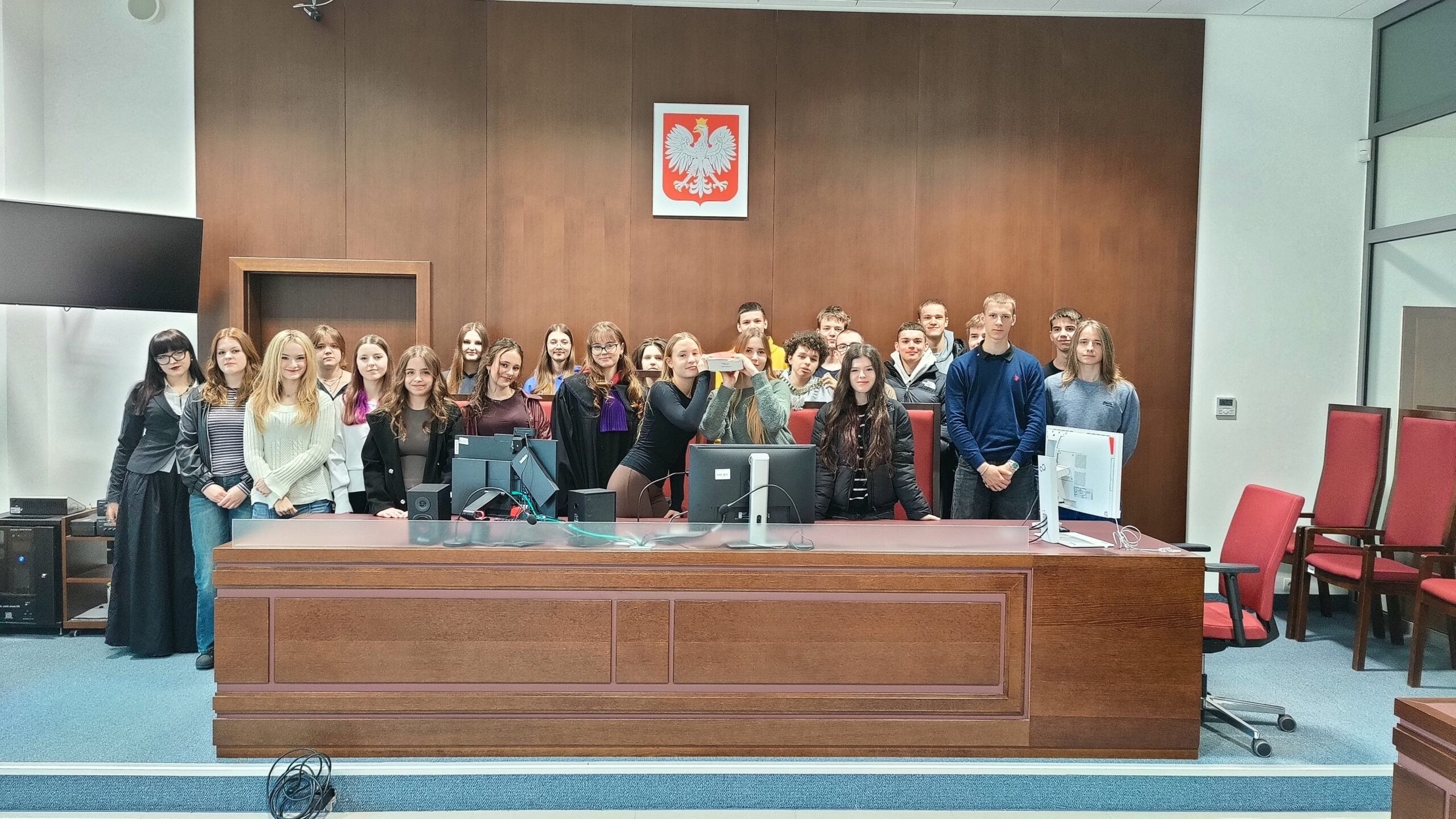 Duża grupa młodych osób stoi za wysokim, drewnianym stołem sędziowskim w sali sądowej. Na ścianie nad nimi widnieje godło Polski – biały orzeł na czerwonym tle. Na stole znajdują się monitory komputerowe, klawiatury i okablowanie. W centrum kilka osób trzyma przedmiot przypominający pudełko lub rekwizyt, prezentując go. Uczestnicy stoją blisko siebie, część uśmiecha się do zdjęcia. Po bokach widać czerwone krzesła oraz elementy wyposażenia sali.