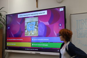 Uczennica stoi przy dużym ekranie z quizem Kahoot. Na ekranie pytanie po polsku i odpowiedzi w kolorowych polach.