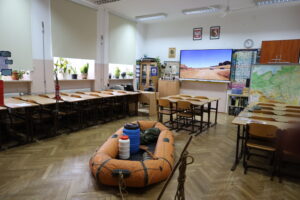 Sala lekcyjna z ławkami, roślinami na parapetach i dużym ekranem. Na podłodze leży pomarańczowy ponton z wyposażeniem.