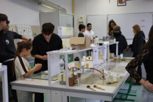 Uczniowie pracują w pracowni chemicznej przy stołach laboratoryjnych z odczynnikami i sprzętem.