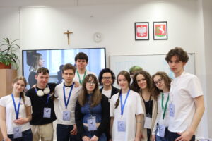 Grupa uczniów i jedna dorosła osoba stoją razem w klasie. Mają identyfikatory na szyi, a w tle widać ekran i tablicę.