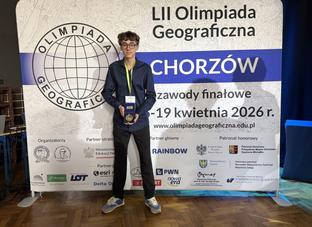 Ten sam chłopak stoi przed dużym banerem „LII Olimpiada Geograficzna – Chorzów”, trzymając medal lub nagrodę i pozując do zdjęcia.