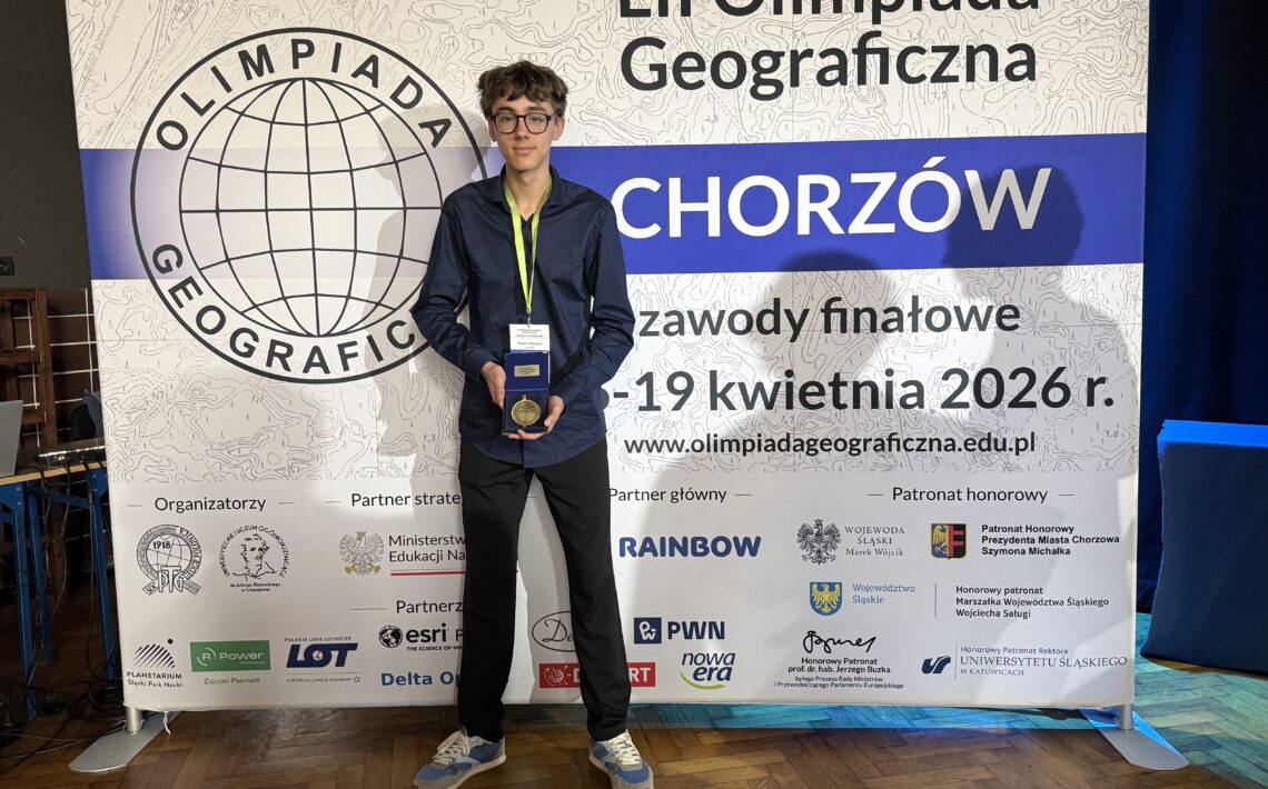 Ten sam chłopak stoi przed dużym banerem „LII Olimpiada Geograficzna – Chorzów”, trzymając medal lub nagrodę i pozując do zdjęcia.
