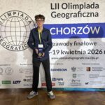 Ten sam chłopak stoi przed dużym banerem „LII Olimpiada Geograficzna – Chorzów”, trzymając medal lub nagrodę i pozując do zdjęcia.