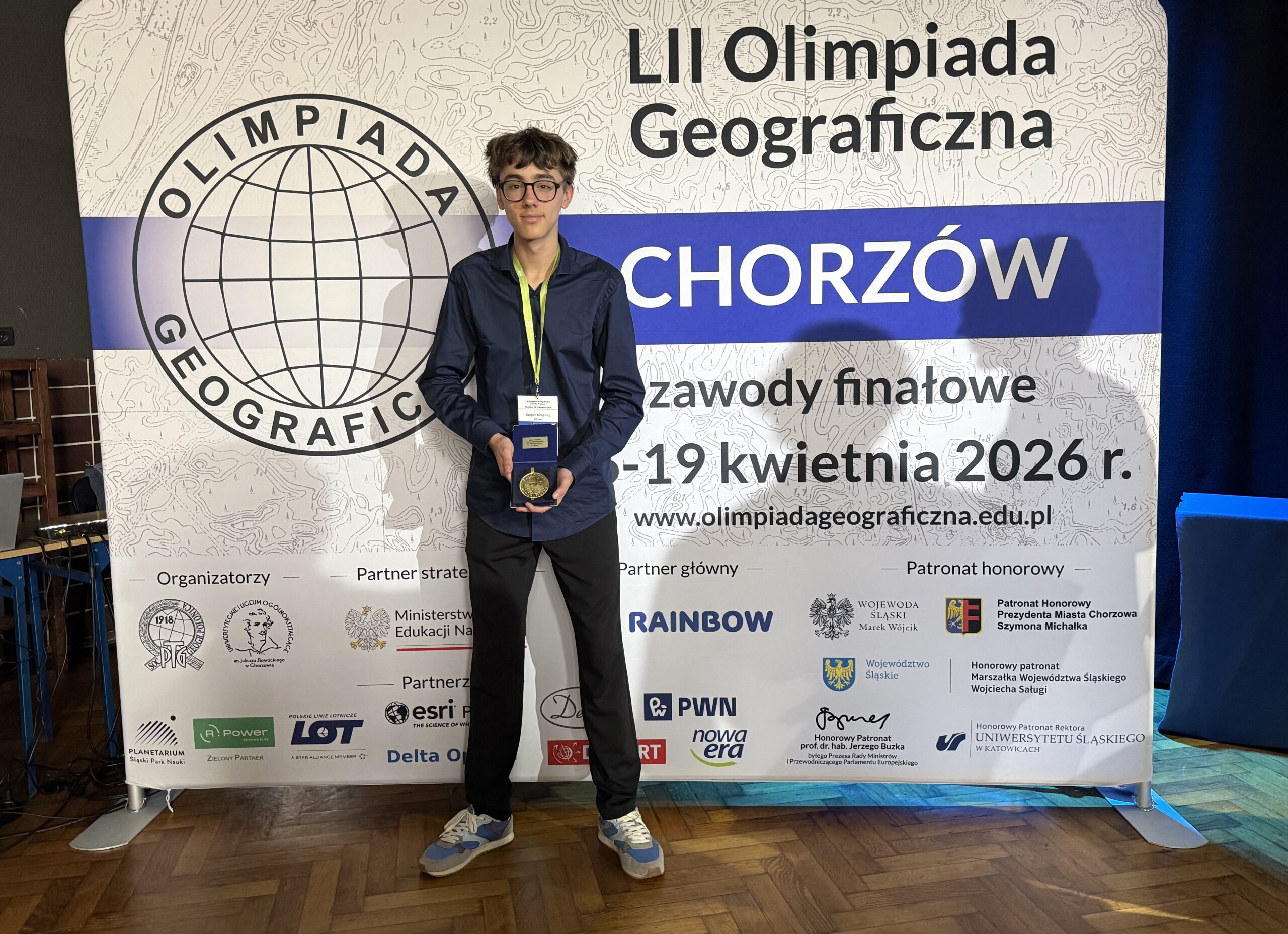 Ten sam chłopak stoi przed dużym banerem „LII Olimpiada Geograficzna – Chorzów”, trzymając medal lub nagrodę i pozując do zdjęcia.