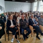 Sala szkolna wypełniona dużą grupą młodzieży siedzącej w rzędach krzeseł. Na pierwszym planie kilka dorosłych osób (kobiety i mężczyźni) siedzi przodem, ubranych elegancko lub półformalnie. W tle gęsto ustawione rzędy uczniów, większość patrzy w jednym kierunku, jakby słuchali wystąpienia. Pomieszczenie jest jasne, z dużymi oknami po prawej stronie i drewnianą podłogą.