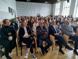 Sala szkolna wypełniona dużą grupą młodzieży siedzącej w rzędach krzeseł. Na pierwszym planie kilka dorosłych osób (kobiety i mężczyźni) siedzi przodem, ubranych elegancko lub półformalnie. W tle gęsto ustawione rzędy uczniów, większość patrzy w jednym kierunku, jakby słuchali wystąpienia. Pomieszczenie jest jasne, z dużymi oknami po prawej stronie i drewnianą podłogą.