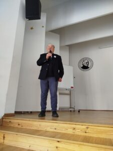 Starszy mężczyzna stoi na niewielkiej scenie i przemawia do publiczności, trzymając mikrofon przy ustach. Ubrany jest w ciemną marynarkę i spodnie. Za nim znajduje się biała ściana z logo szkoły oraz tablica na stojaku. Scena ma kilka drewnianych stopni, a nad nią widoczny jest głośnik.