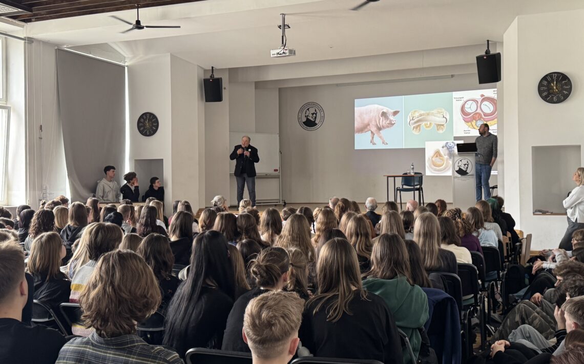 Sala wykładowa wypełniona siedzącymi uczestnikami (głównie młodzieżą) skierowanymi w stronę sceny. Z przodu stoi dwóch mężczyzn prowadzących prezentację; na ekranie wyświetlone są slajdy z ilustracjami (m.in. świnia i schematy anatomiczne). W pomieszczeniu widać zegary ścienne, głośniki, wentylatory sufitowe oraz projektor zamocowany pod sufitem.