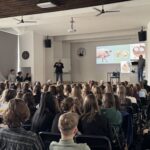 Sala wykładowa wypełniona siedzącymi uczestnikami (głównie młodzieżą) skierowanymi w stronę sceny. Z przodu stoi dwóch mężczyzn prowadzących prezentację; na ekranie wyświetlone są slajdy z ilustracjami (m.in. świnia i schematy anatomiczne). W pomieszczeniu widać zegary ścienne, głośniki, wentylatory sufitowe oraz projektor zamocowany pod sufitem.