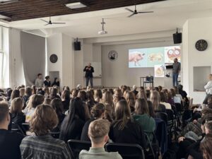Sala wykładowa wypełniona siedzącymi uczestnikami (głównie młodzieżą) skierowanymi w stronę sceny. Z przodu stoi dwóch mężczyzn prowadzących prezentację; na ekranie wyświetlone są slajdy z ilustracjami (m.in. świnia i schematy anatomiczne). W pomieszczeniu widać zegary ścienne, głośniki, wentylatory sufitowe oraz projektor zamocowany pod sufitem.