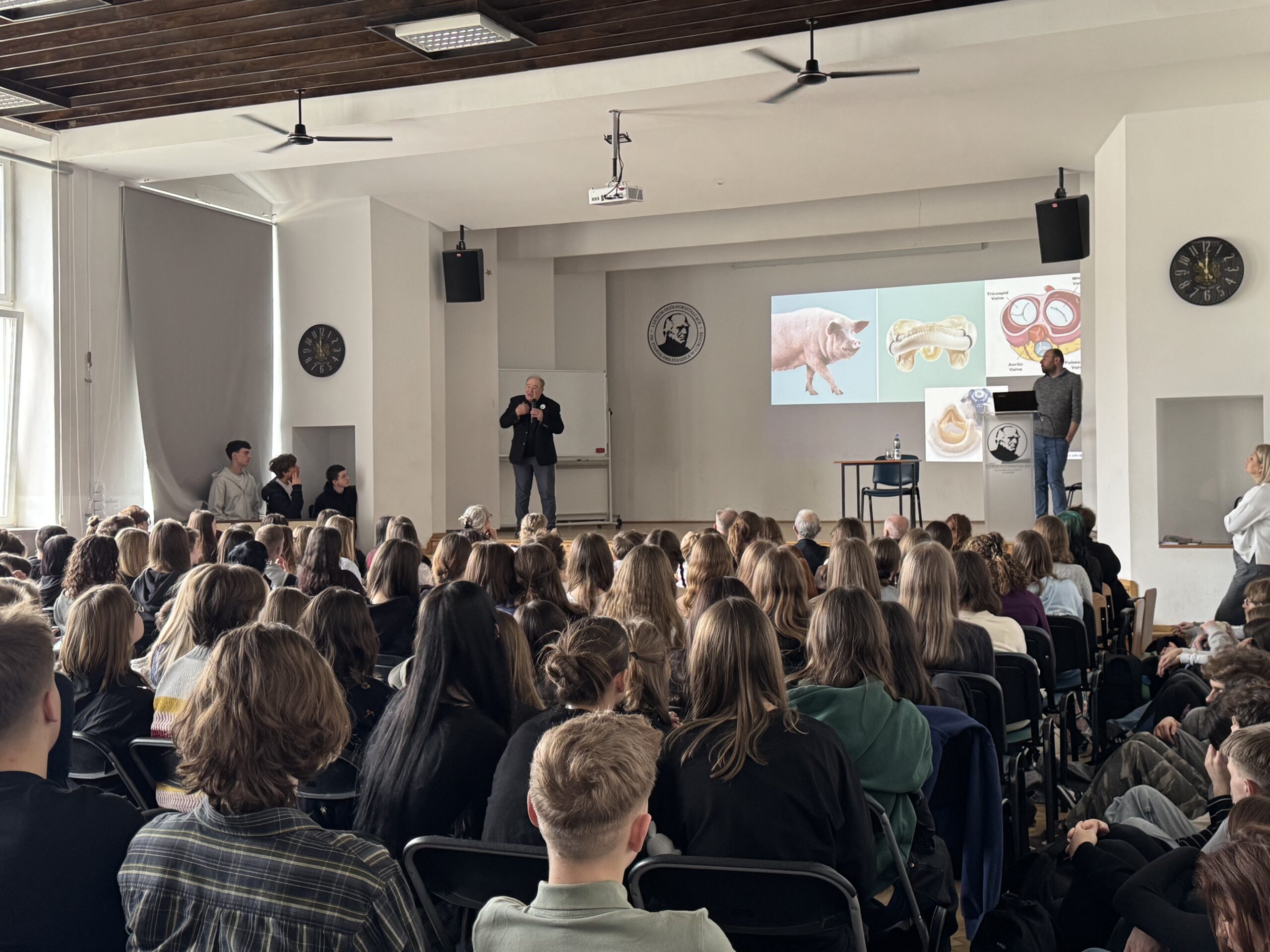 Sala wykładowa wypełniona siedzącymi uczestnikami (głównie młodzieżą) skierowanymi w stronę sceny. Z przodu stoi dwóch mężczyzn prowadzących prezentację; na ekranie wyświetlone są slajdy z ilustracjami (m.in. świnia i schematy anatomiczne). W pomieszczeniu widać zegary ścienne, głośniki, wentylatory sufitowe oraz projektor zamocowany pod sufitem.