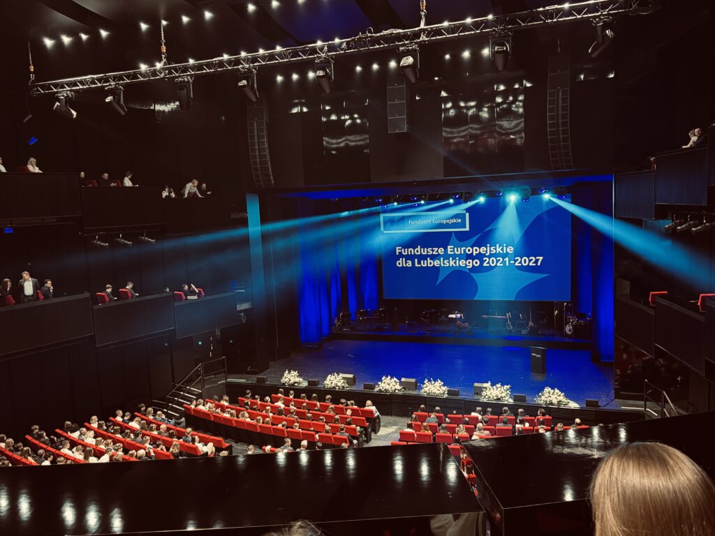 Szeroki widok całej sali widowiskowej z czerwonymi fotelami i publicznością. Na scenie pustej, przygotowanej do wydarzenia, widoczny jest duży ekran z napisem: „Fundusze Europejskie dla Lubelskiego 2021–2027”. Oświetlenie w odcieniach niebieskiego, reflektory skierowane na scenę.