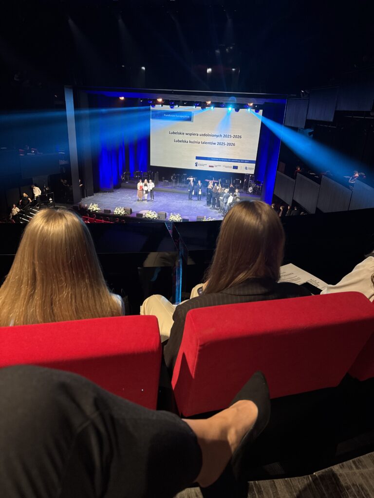 Widok z balkonu na scenę w dużej, nowoczesnej sali teatralnej. Na pierwszym planie czerwone fotele i dwie osoby siedzące tyłem. Na scenie grupa ludzi odbiera nagrody, a nad nimi duży ekran z napisem: „Lubelskie wspiera uzdolnionych 2025–2026. Lubelska kuźnia talentów 2025–2026”. Niebieskie światła sceniczne skierowane są na uczestników.