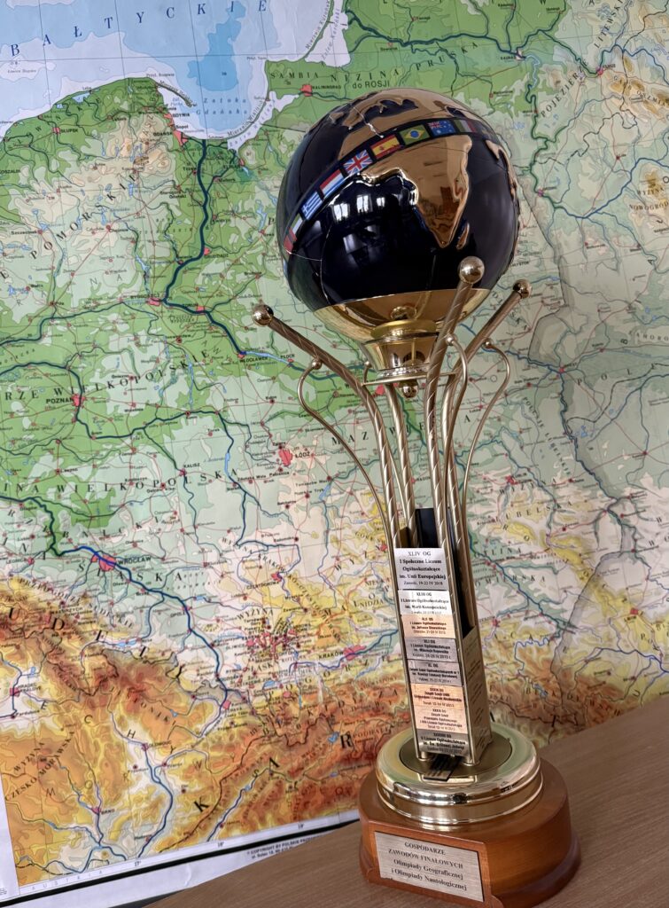 Okazały puchar z kulą ziemską na szczycie stoi na tle szczegółowej mapy Polski; na podstawie widoczna jest tabliczka z opisem nagrody.