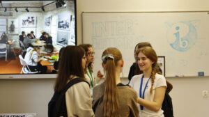 Kilka uczennic rozmawia w klasie przed tablicą z napisem „International Baccalaureate (IB)”. Na ekranie obok wyświetlone zdjęcie uczniów przy pracy.