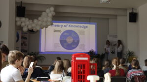 Sala z prezentacją – uczniowie siedzą przodem do ekranu z napisem „Theory of Knowledge”. Na środku stoi dekoracyjna czerwona budka telefoniczna, a przy podium dwie osoby prowadzą wystąpienie.