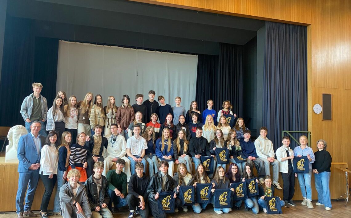 Duża grupa młodzieży i kilku dorosłych (około 40–50 osób) ustawiona w trzech rzędach na scenie w sali z drewnianymi ścianami. Część osób siedzi, część stoi. Kilka osób trzyma granatowe torby z żółtym nadrukiem. W tle jasna kurtyna. Po prawej zegar na ścianie. Wszyscy pozują do wspólnego zdjęcia.