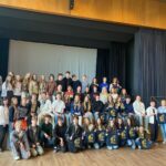 Duża grupa młodzieży i kilku dorosłych (około 40–50 osób) ustawiona w trzech rzędach na scenie w sali z drewnianymi ścianami. Część osób siedzi, część stoi. Kilka osób trzyma granatowe torby z żółtym nadrukiem. W tle jasna kurtyna. Po prawej zegar na ścianie. Wszyscy pozują do wspólnego zdjęcia.