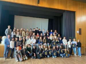 Duża grupa młodzieży i kilku dorosłych (około 40–50 osób) ustawiona w trzech rzędach na scenie w sali z drewnianymi ścianami. Część osób siedzi, część stoi. Kilka osób trzyma granatowe torby z żółtym nadrukiem. W tle jasna kurtyna. Po prawej zegar na ścianie. Wszyscy pozują do wspólnego zdjęcia.