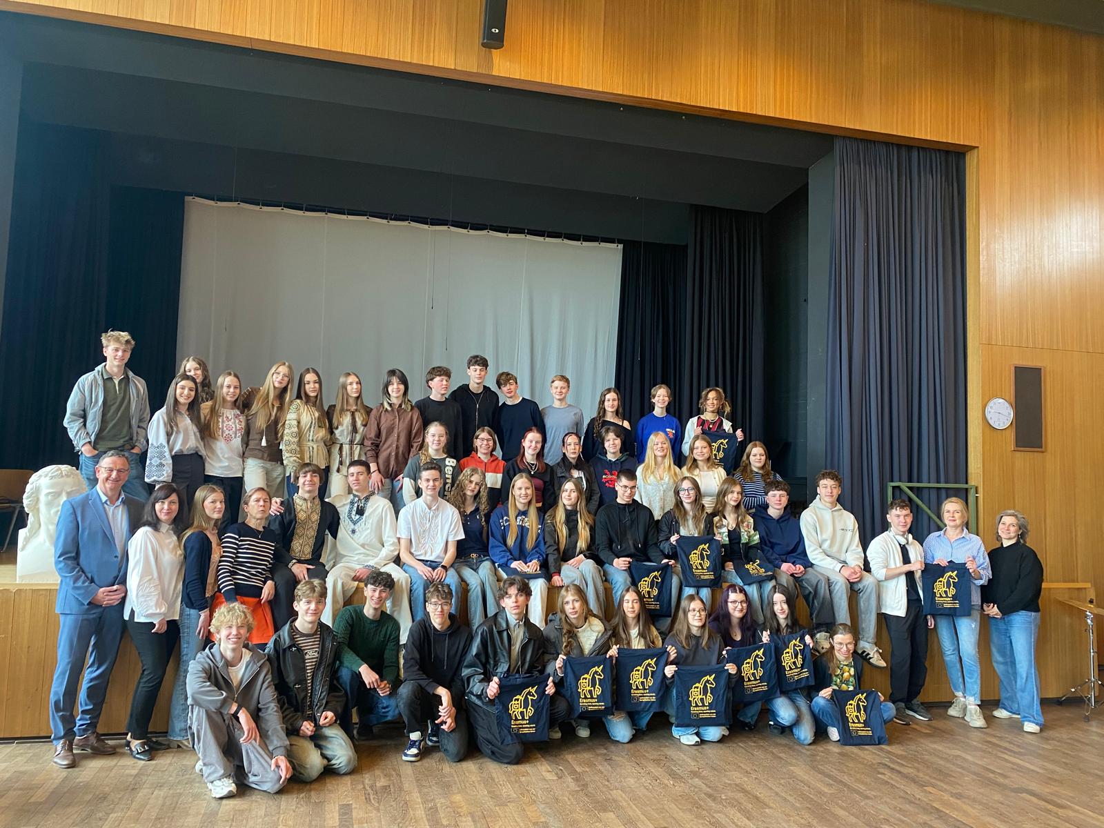 Duża grupa młodzieży i kilku dorosłych (około 40–50 osób) ustawiona w trzech rzędach na scenie w sali z drewnianymi ścianami. Część osób siedzi, część stoi. Kilka osób trzyma granatowe torby z żółtym nadrukiem. W tle jasna kurtyna. Po prawej zegar na ścianie. Wszyscy pozują do wspólnego zdjęcia.