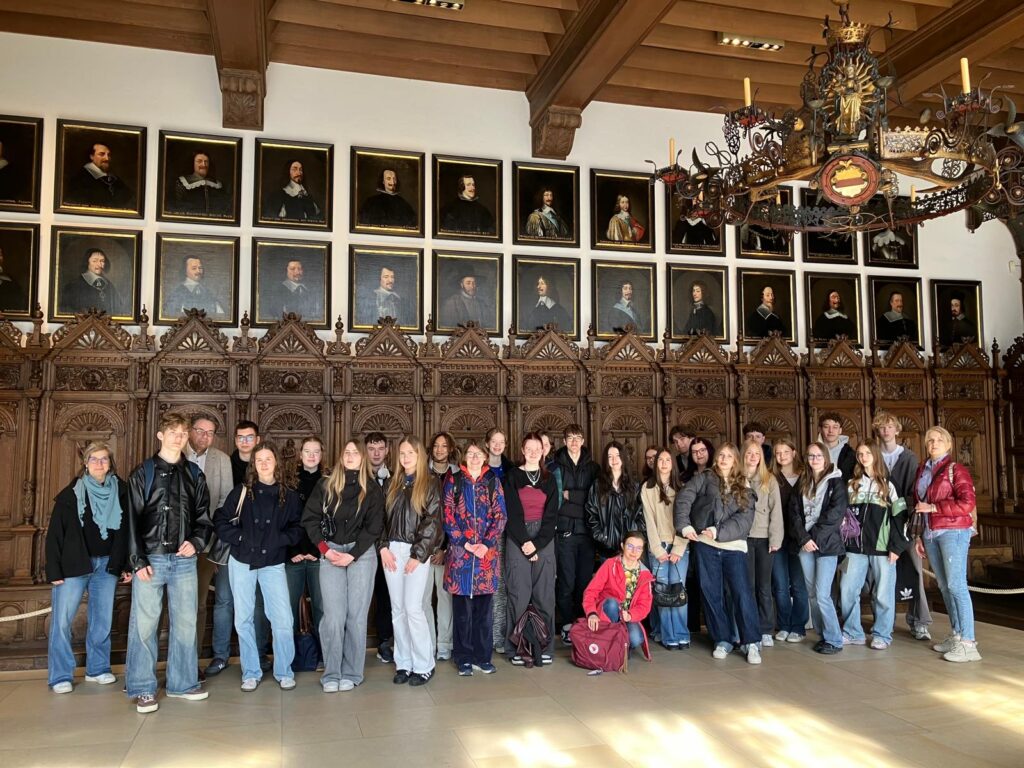 Grupa młodzieży i opiekunów (około 25–30 osób) stoi w bogato zdobionej sali z drewnianym stropem. Za nimi rząd rzeźbionych drewnianych stall i galeria portretów historycznych postaci w złotych ramach. Nad grupą duży ozdobny żyrandol. Osoby stoją blisko siebie, patrzą w stronę aparatu.