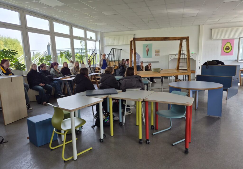Nowoczesna sala lekcyjna z dużymi oknami po lewej stronie. Kilkanaście osób siedzi przy stołach i na ławkach ustawionych w półokręgu, słuchając prowadzącej stojącej przy stole demonstracyjnym. Meble są kolorowe (jasne blaty, nogi w różnych kolorach). Na ścianie grafiki (m.in. lody i awokado). Atmosfera zajęć warsztatowych.
