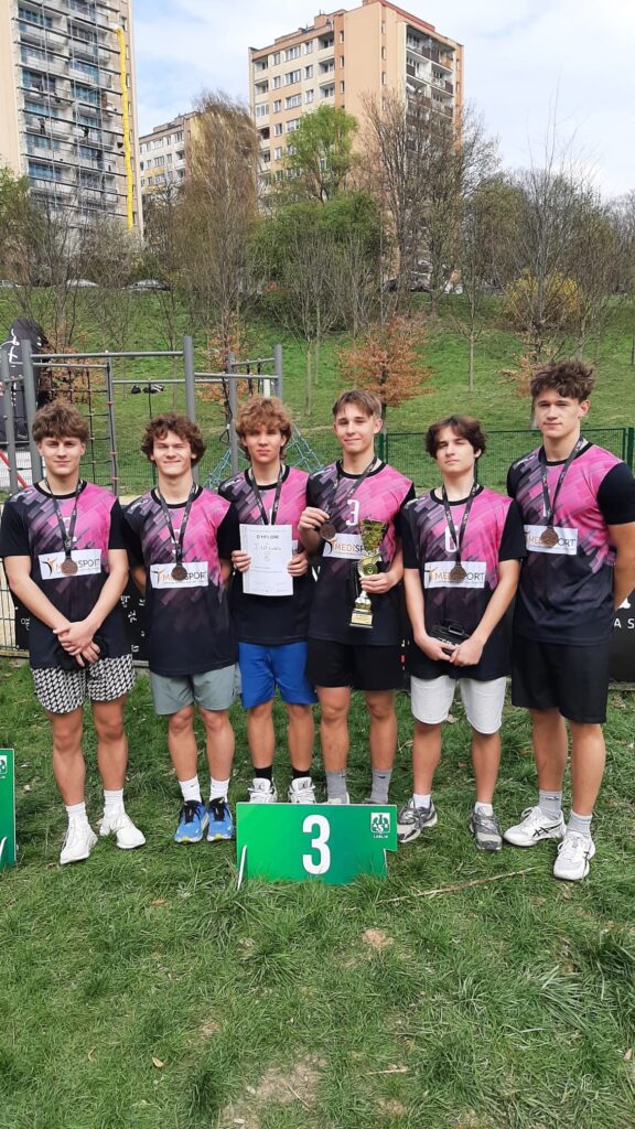 Sześciu nastoletnich chłopców stoi obok siebie na trawie przed blokami i placem do ćwiczeń. Mają czarno-różowe stroje sportowe i medale na szyi. Jeden trzyma puchar, inny dyplom. Przed nimi zielona tabliczka z dużą cyfrą „3”.