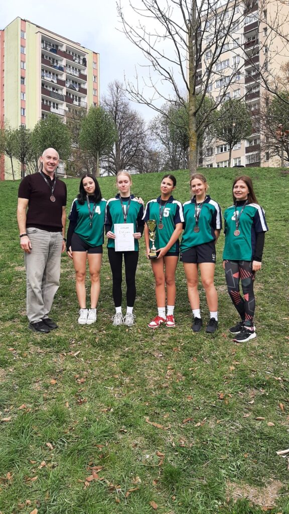 Grupa sześciu osób stoi na trawie w parku między blokami mieszkalnymi. Po lewej dorosły mężczyzna, obok pięć nastolatek w zielonych koszulkach sportowych i czarnych spodenkach/legginsach. Wszystkie mają medale na szyi; jedna trzyma dyplom, druga puchar. W tle drzewa bez liści i wysokie budynki.