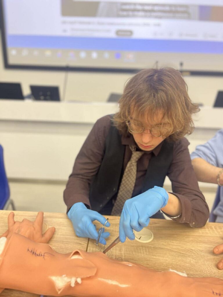 Młody mężczyzna w rękawiczkach wykonuje ćwiczenie medyczne na modelu ręki przy stole, w tle widoczna sala wykładowa.