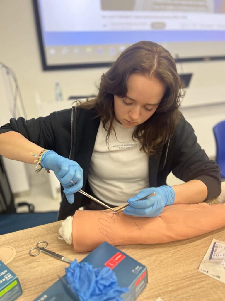 Młoda kobieta w rękawiczkach precyzyjnie pracuje narzędziami na modelu ręki podczas zajęć praktycznych w sali dydaktycznej.