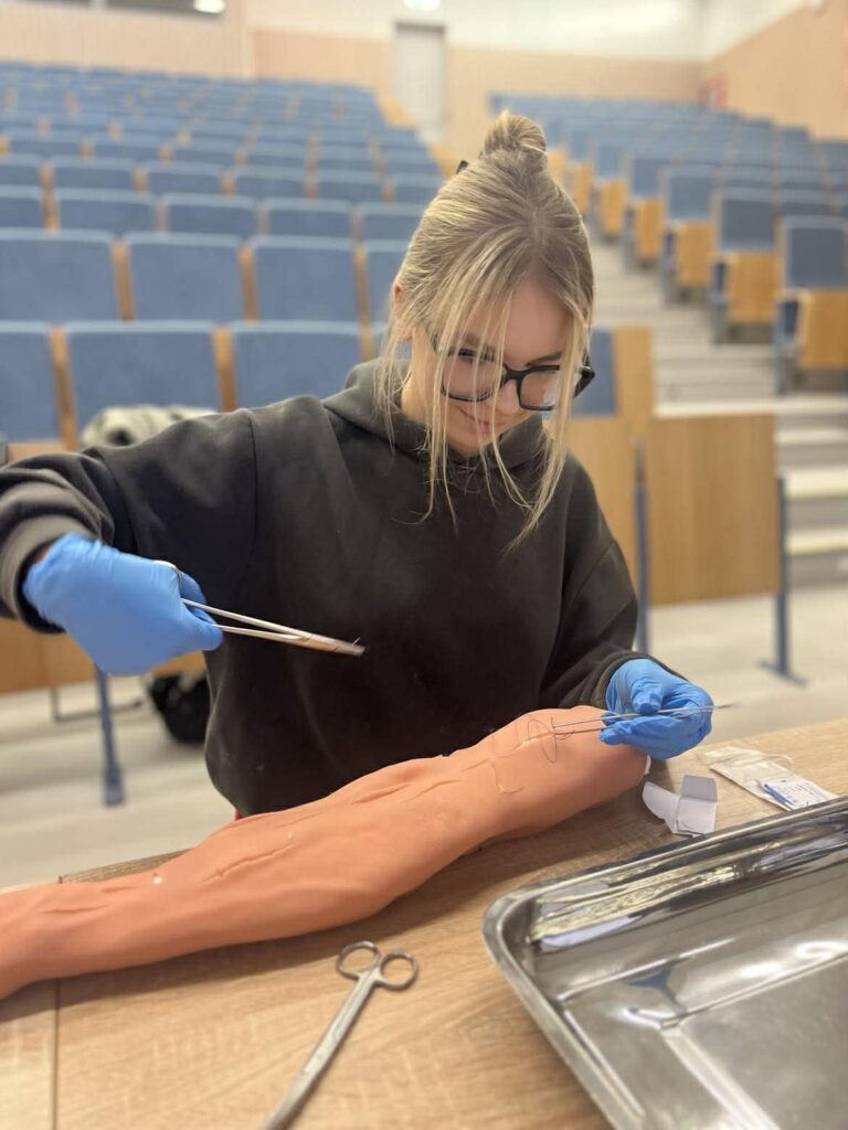 Młoda kobieta w okularach i rękawiczkach wykonuje ćwiczenie medyczne na modelu ręki, używając narzędzi chirurgicznych przy stole.