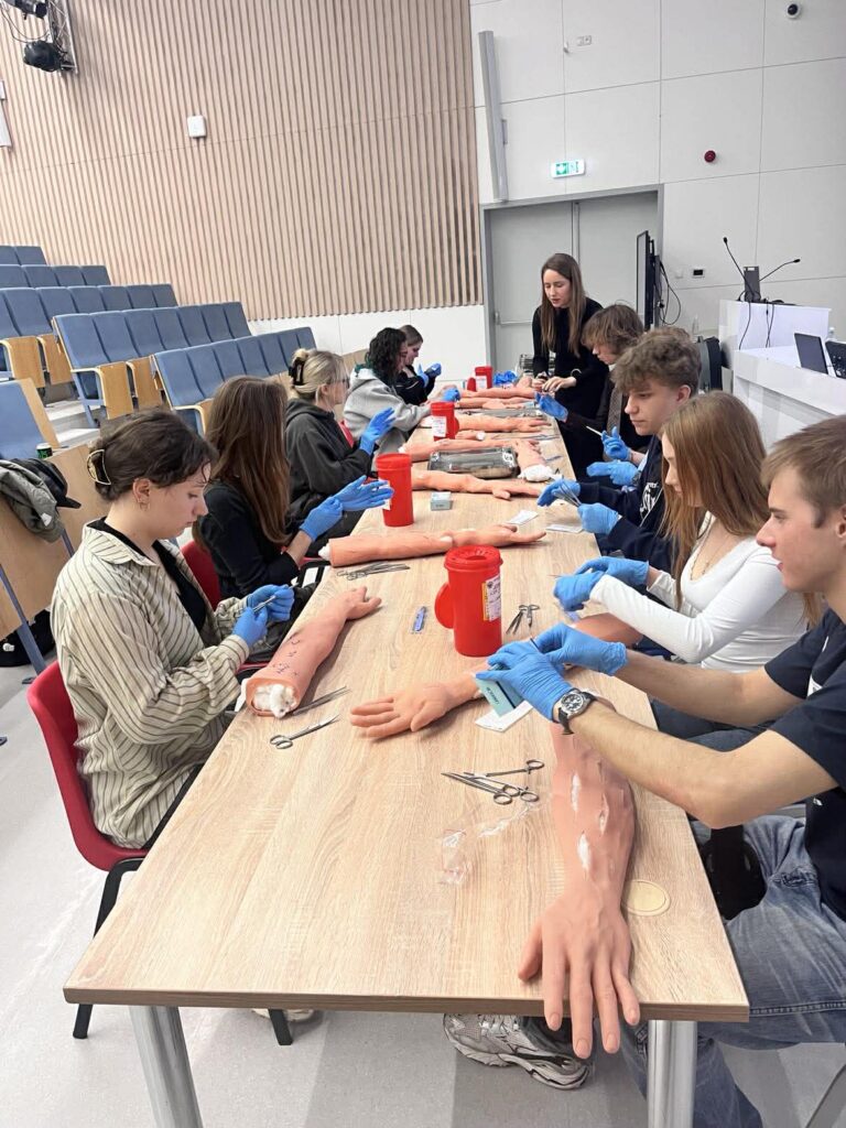 Studenci siedzą przy stole i ćwiczą procedury medyczne na realistycznych modelach rąk, używając narzędzi i rękawiczek ochronnych. W tle sala wykładowa.