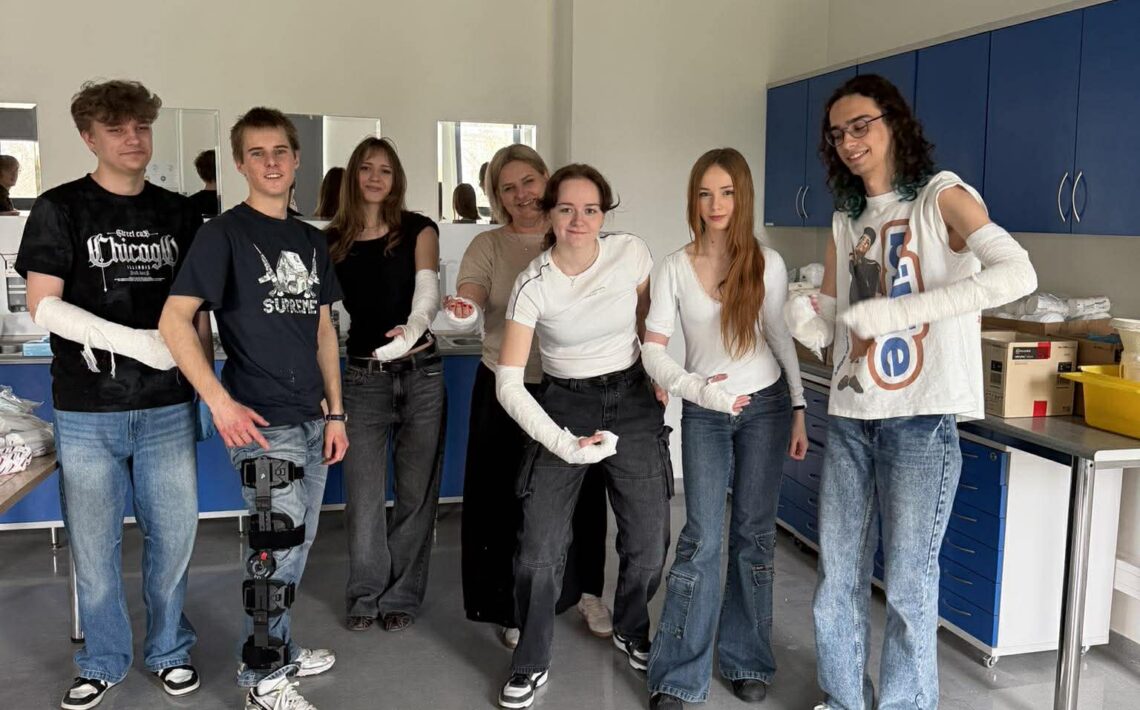 Grupa siedmiu młodych osób stoi w jasnej sali medycznej; większość ma zabandażowane ręce. W tle szafki i lustra, atmosfera ćwiczeń praktycznych.