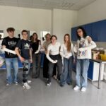 Grupa siedmiu młodych osób stoi w jasnej sali medycznej; większość ma zabandażowane ręce. W tle szafki i lustra, atmosfera ćwiczeń praktycznych.