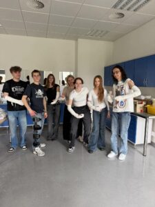 Grupa siedmiu młodych osób stoi w jasnej sali medycznej; większość ma zabandażowane ręce. W tle szafki i lustra, atmosfera ćwiczeń praktycznych.