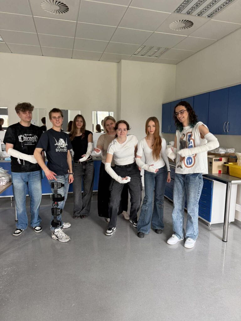 Grupa siedmiu młodych osób stoi w jasnej sali medycznej; większość ma zabandażowane ręce. W tle szafki i lustra, atmosfera ćwiczeń praktycznych.