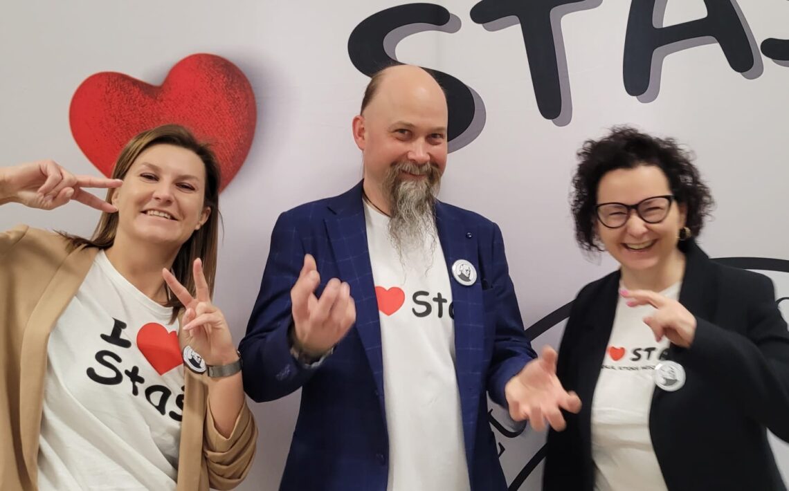Trzy dorosłe osoby pozują na tle napisu „STAŚ”. Dwie kobiety robią gest „V” palcami, a mężczyzna stoi pośrodku w marynarce i koszulce z sercem.