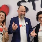 Trzy dorosłe osoby pozują na tle napisu „STAŚ”. Dwie kobiety robią gest „V” palcami, a mężczyzna stoi pośrodku w marynarce i koszulce z sercem.