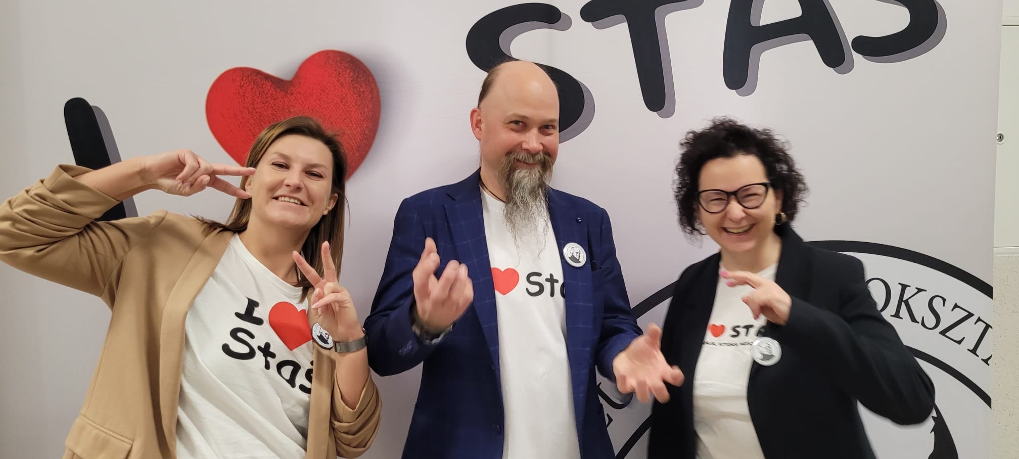 Trzy dorosłe osoby pozują na tle napisu „STAŚ”. Dwie kobiety robią gest „V” palcami, a mężczyzna stoi pośrodku w marynarce i koszulce z sercem.