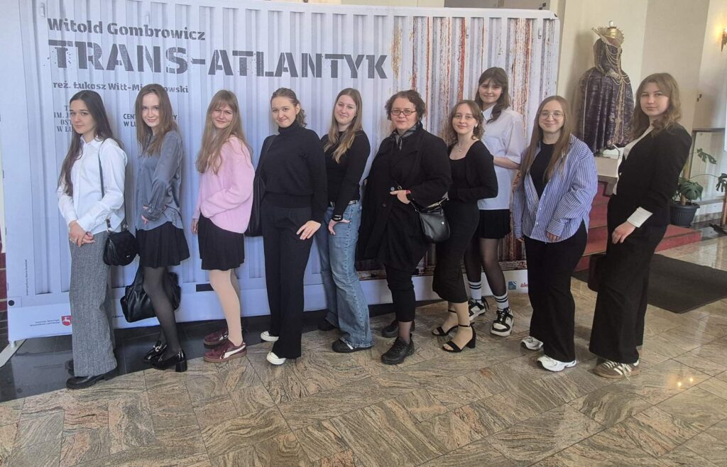 Grupa młodych kobiet stoi w rzędzie przed dużym plakatem „Trans-Atlantyk” Witolda Gombrowicza; są ustawione bokiem, uśmiechnięte, w półformalnych ubraniach.