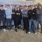 Grupa młodych mężczyzn z nauczycielką pośrodku stoi przed tym samym plakatem; większość ma skrzyżowane ręce i patrzy w stronę aparatu.